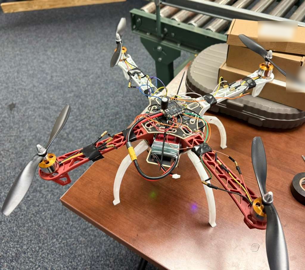 A four-rotor drone with a red and white frame, black propellers and exposed wiring sits on a workbench, its flight controller board mounted at the center.