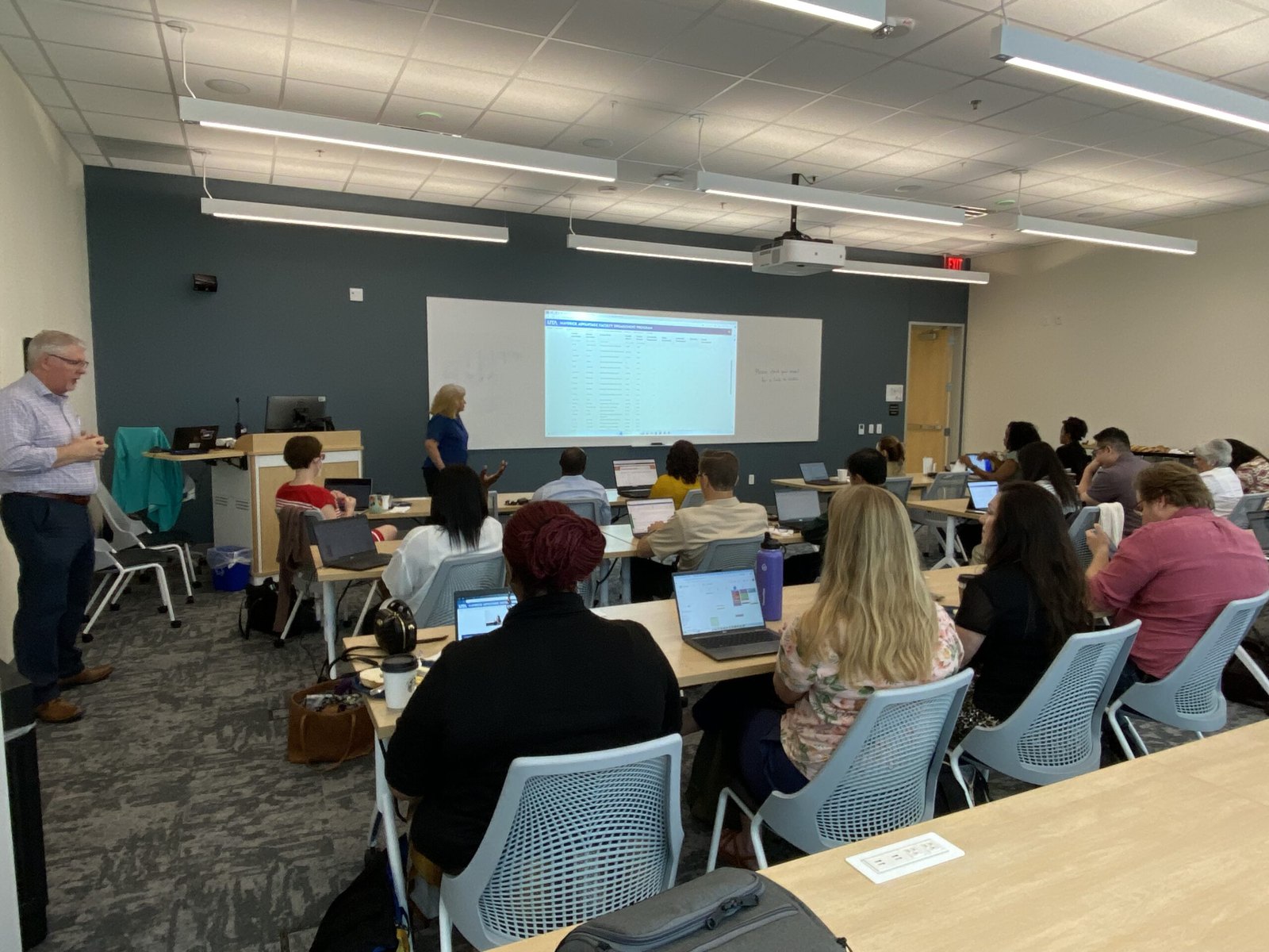 Faculty members seated at laptops in a UTA classroom during a sustainability teaching workshop, with a presenter at the front of the room and a web page projected on the screen.