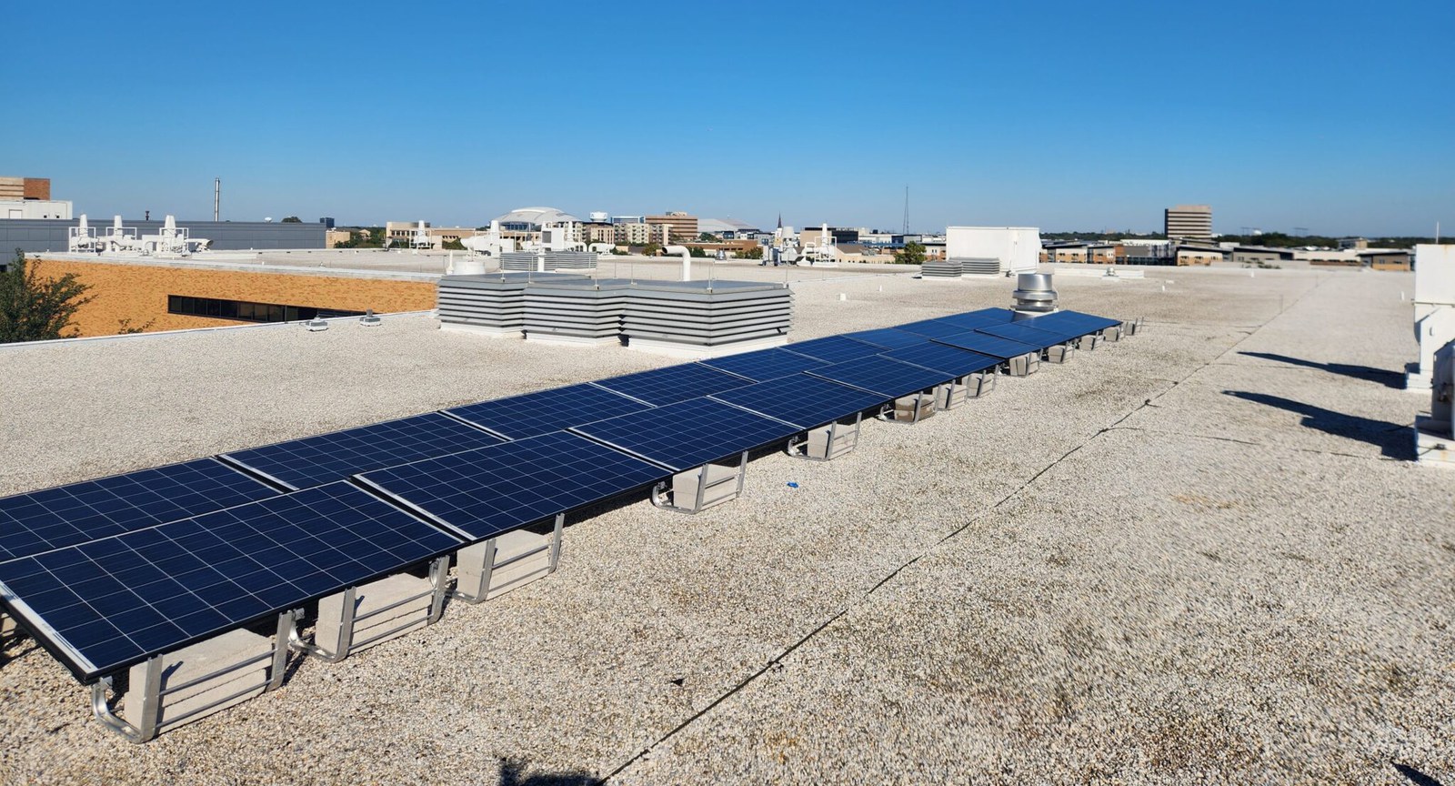 A long row of solar panels on ballasted racks across a flat gravel roof, with low campus buildings and the Arlington skyline under a clear blue sky.