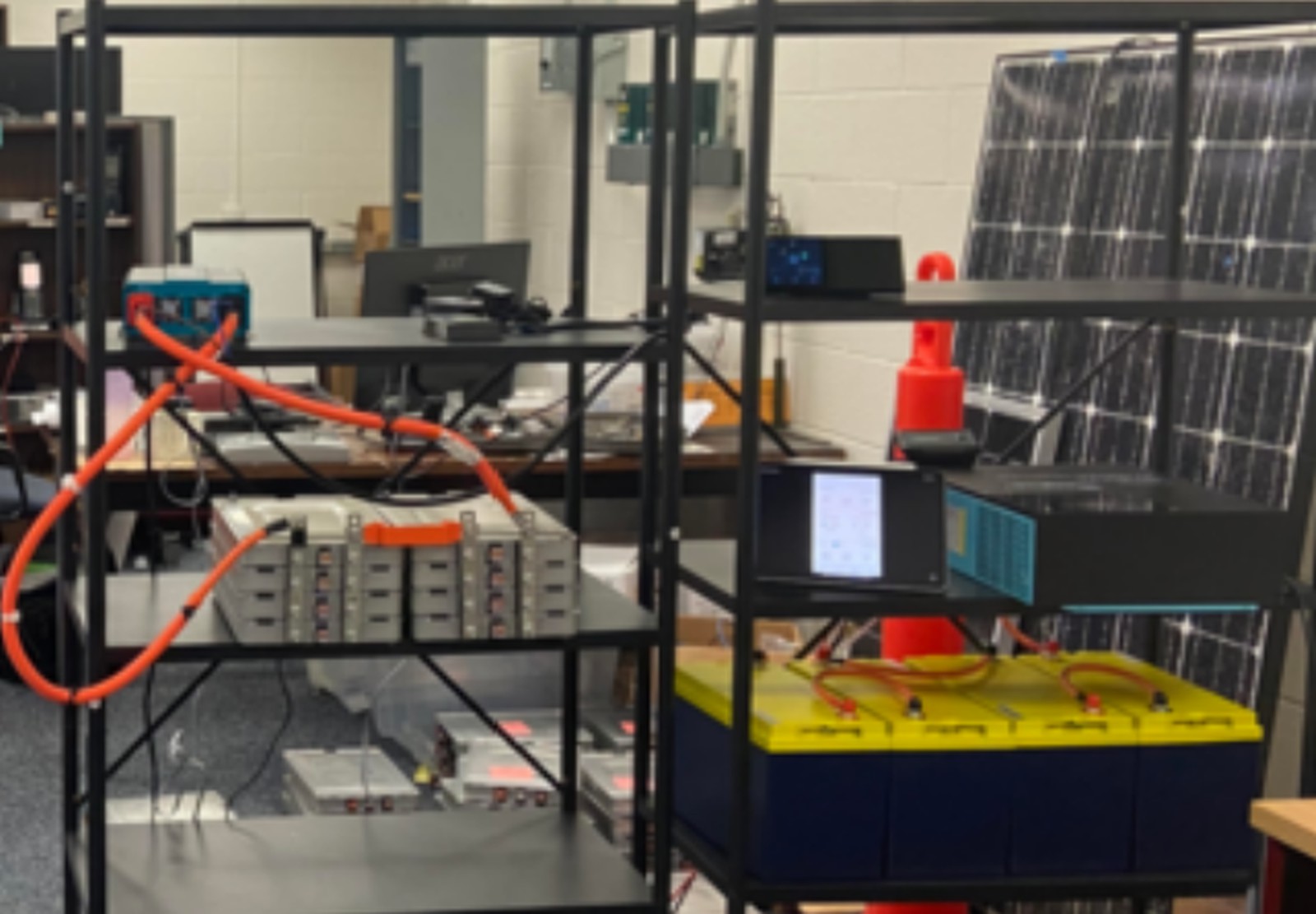 Two black metal shelving units: the left holds grey battery modules joined by orange high-voltage cable and a bench instrument; the right holds instruments, a yellow-topped battery bank and solar panels leaning behind.